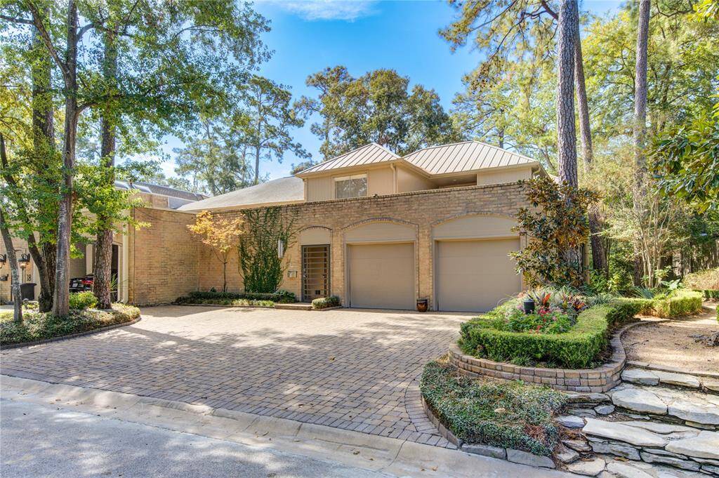Charming, two-story brick home with a metal roof nestled among mature trees. It has a tidy, landscaped front yard with a dual garage and a stone pathway. The driveway is paved with bricks, offering a welcoming entrance.