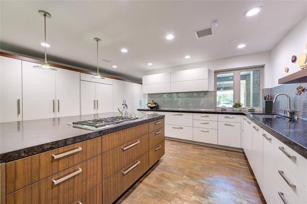 This recent modern kitchen features sleek white cabinetry, a large central island with a gas cooktop, and pendant lighting. It offers ample counter space, a mosaic tile backsplash, and a bright, open atmosphere with plenty of natural light.