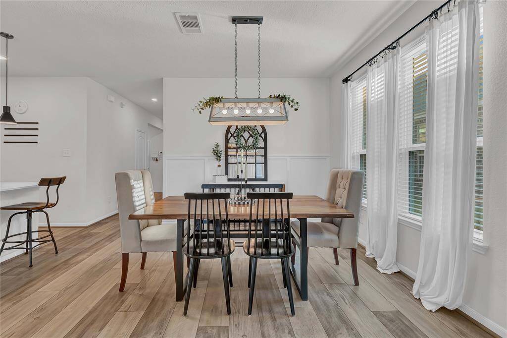 This dining area features a modern, cozy design with a wooden table, mixed seating, and a stylish overhead light fixture. Natural light floods through large windows with sheer curtains, complementing the light wood flooring.