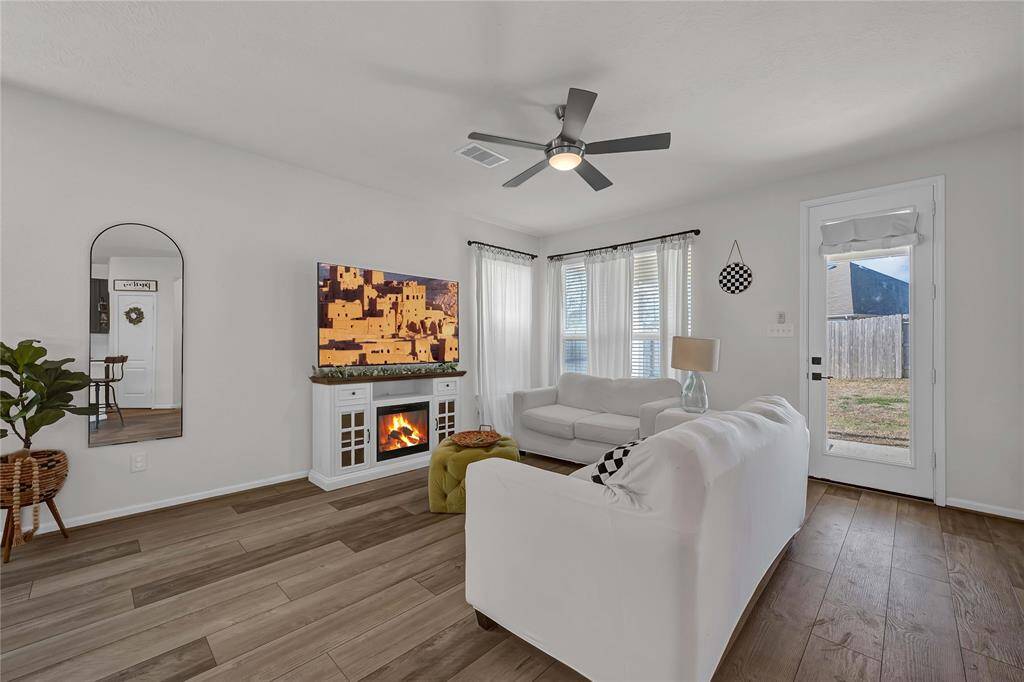 Bright and welcoming living room featuring hardwood floors, white walls, and a ceiling fan. The space is furnished with light-colored sofas, a modern media console with a TV, and a cozy electric fireplace. Large windows and a glass door provide ample natural light and access to the backyard.