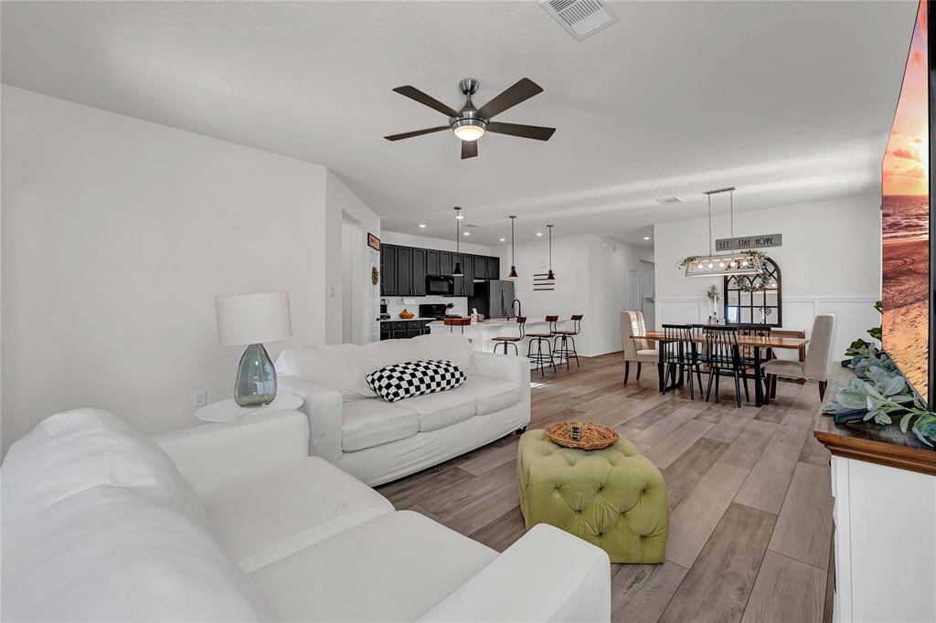 This open-concept living space features a cozy seating area with a modern ceiling fan, a stylish dining area, and a sleek kitchen with dark cabinetry and pendant lighting. The room is bright and welcoming, with wood-look flooring and contemporary decor.