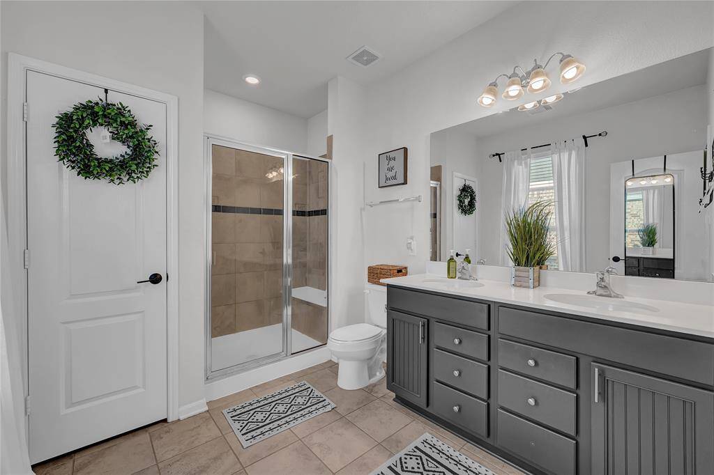 This bathroom features a modern double-sink vanity with ample storage, a glass-enclosed shower with tile accents, and neutral tones. Natural light streams in through a window with sheer curtains, creating a bright and inviting space.