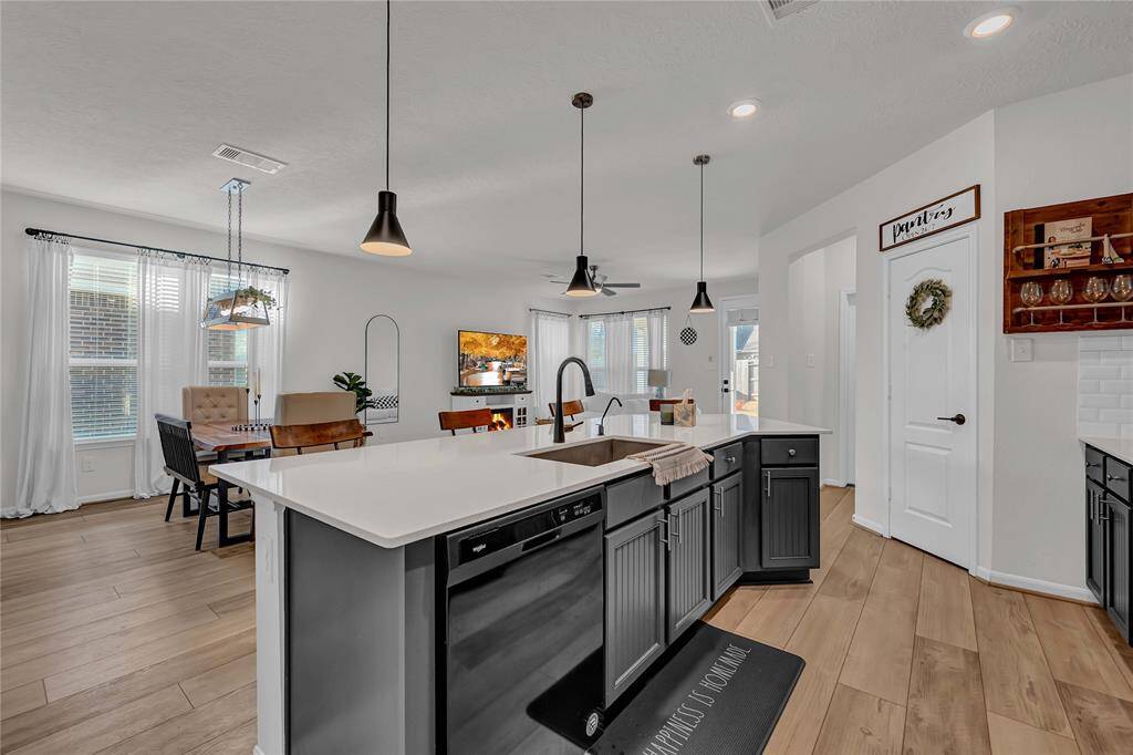 This open-concept kitchen and dining area features modern pendant lighting, a spacious island with a sink, and sleek black cabinetry. The light wood flooring and large windows create a bright, inviting atmosphere, perfect for entertaining.