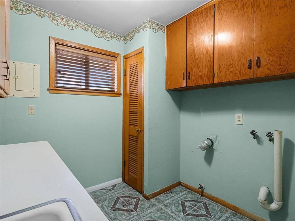 In-home utility/laundry room with light aqua blue walls & decorative trim, features wood cabinetry and louvered wooden door to storage. Washer/dryer connections, counter with sink, natural light from a window with blinds complete this pretty and practical space.  Cabinets are also tile lined !