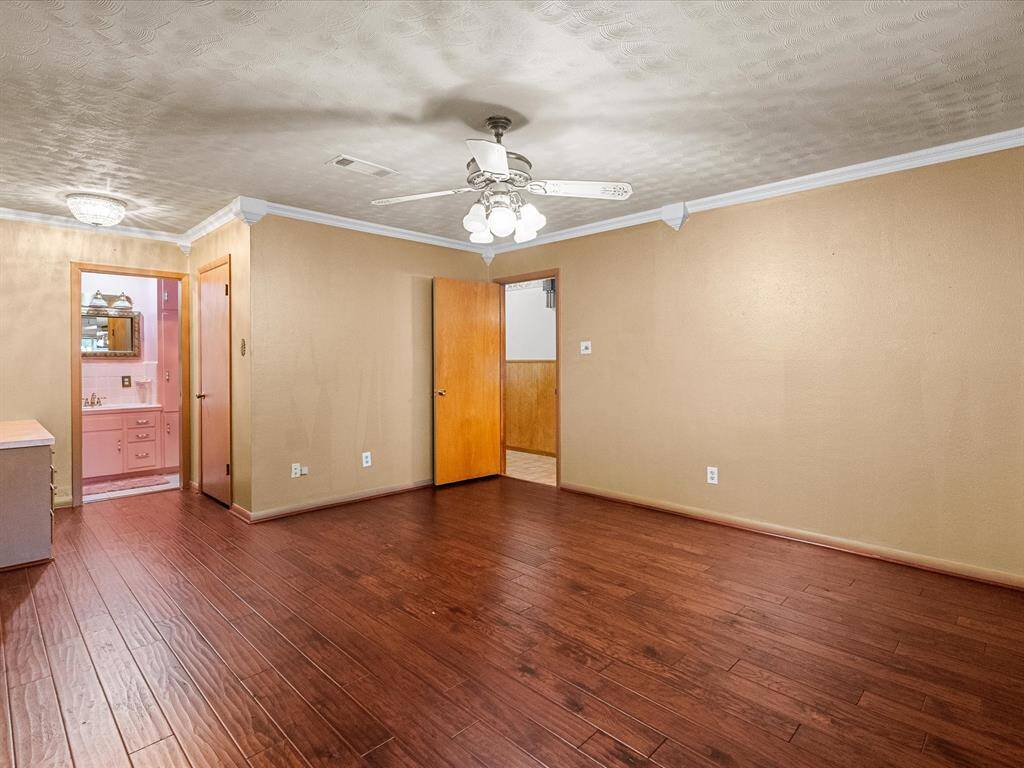 Primary bedroom features rich wooden flooring, ceiling fan, crown molding & neutral wall tones. Adjoining bathroom with pink cabinetry, adds a touch of character. Corner custom built-in vanity w/drawers on both side; lock-n-key on left & right bottom drawers.