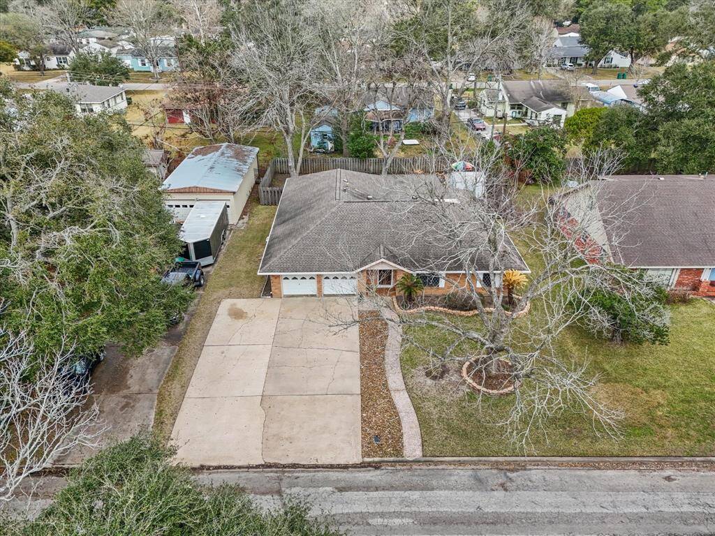 Aerial view showcases a single-story home with a spacious driveway and attached garage. The house is surrounded by mature trees and a fenced backyard, offering privacy in an overall well-maintained neighborhood.