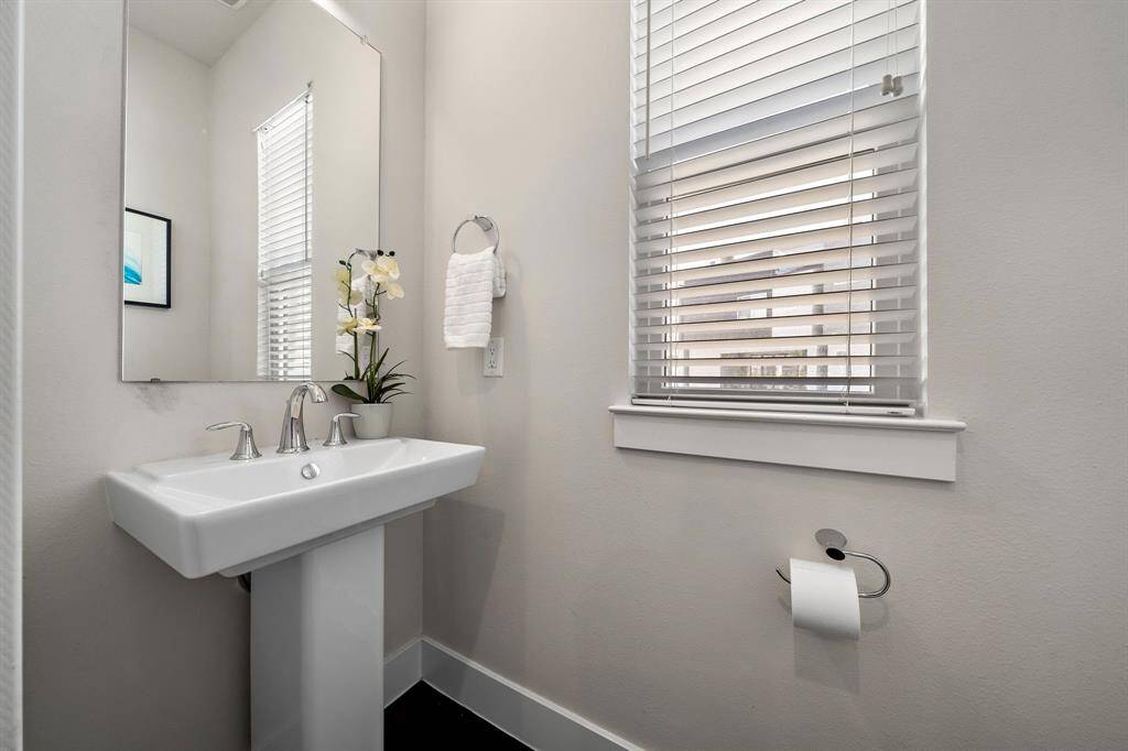 Contemporary half bath features a sleek pedestal sink, framed by a large mirror and modern fixtures. Natural light filters through the window, enhancing the clean lines and minimalistic design, making this space both functional and stylish.