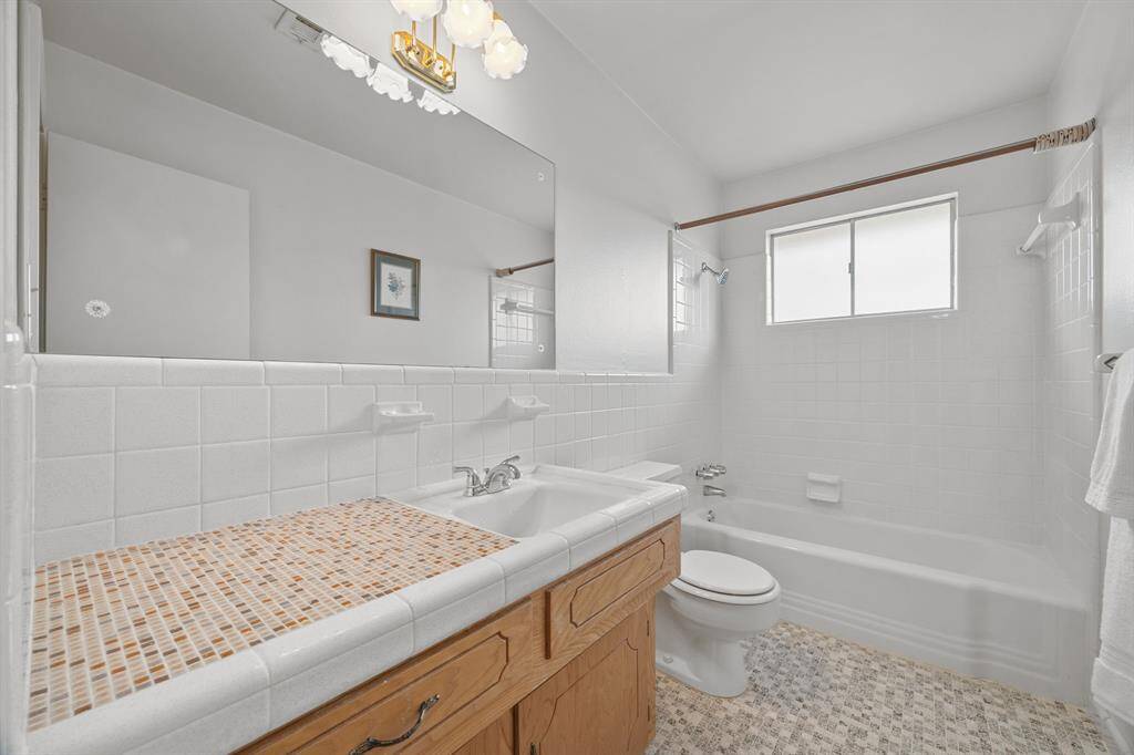 This Bathroom features a clean, bright design with a large mirror, a single sink vanity with mosaic countertop, and a window that lets in natural light. The full bathroom has a Bathtub with a Tub/Shower combination with white tile accents.