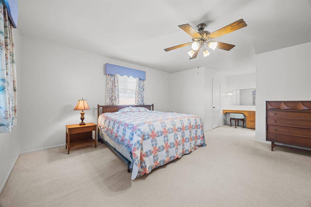 This spacious Primary Bedroom features neutral carpeting, a ceiling fan, and natural light from a window with floral curtains. Primary Bathroom has a well lite Vanity Area, creating a cozy and functional space.