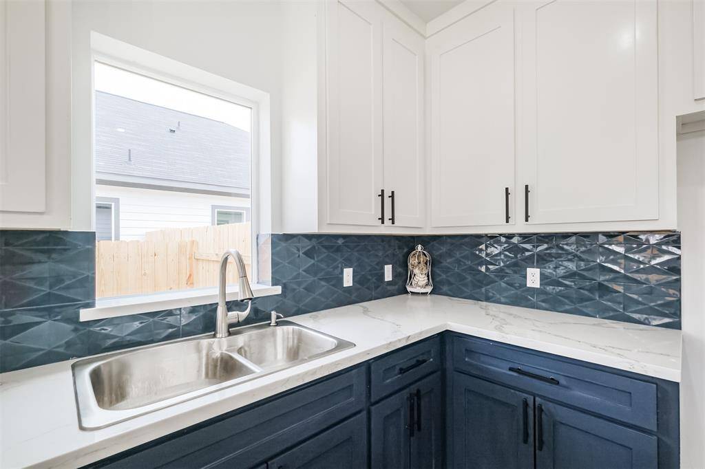 This modern kitchen features sleek white and navy cabinetry with black hardware, a stylish geometric backsplash, and elegant marble countertops. The space includes stainless steel appliances, a spacious island with contemporary lighting, and dark hardwood flooring, offering a sophisticated and functional design.
