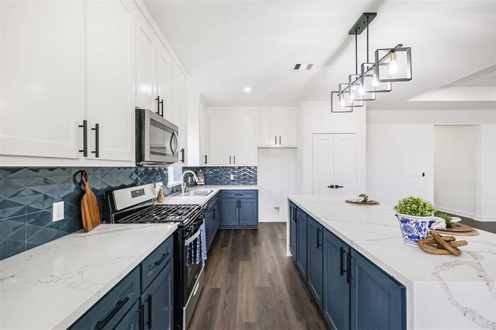 This photo showcases a spacious, open-concept kitchen and living area with modern finishes. The kitchen features a large island, dark cabinetry, and stainless steel appliances, complemented by sleek lighting fixtures. The room is bright with recessed lighting and a ceiling fan, and the flooring is a rich wood tone, offering a clean and contemporary feel.