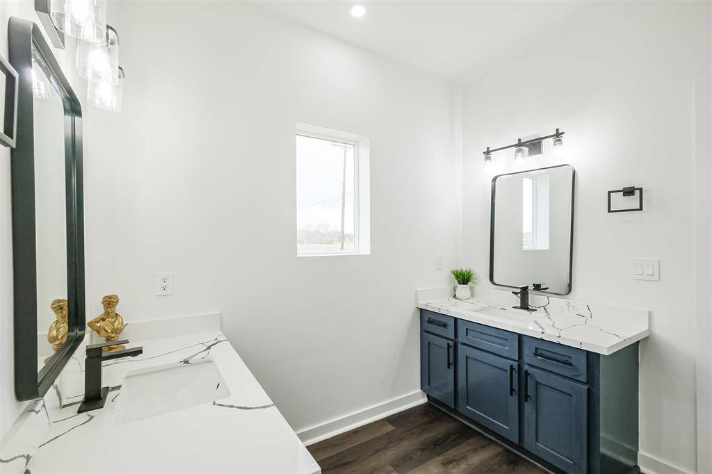 This is a modern bathroom featuring dual vanities with sleek white countertops and dark blue cabinets. It has a minimalist design with a large mirror, wood flooring, recessed lighting, and a small window allowing natural light.