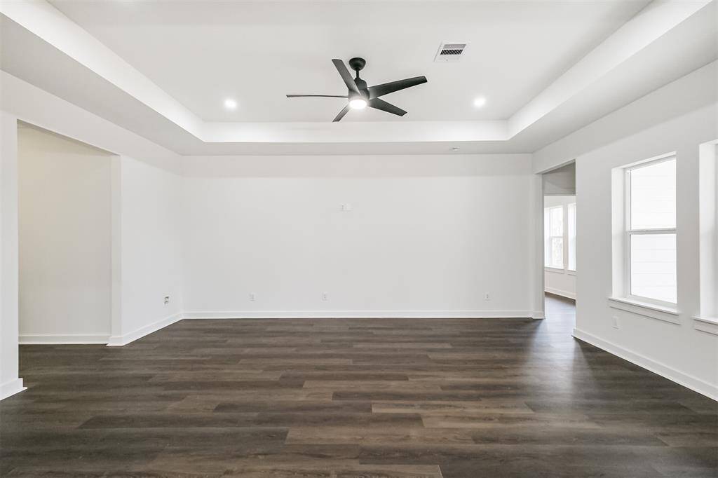 This photo showcases a spacious, modern living area with dark wood flooring, a ceiling fan, and ample natural light from large windows. The room flows into a kitchen area with a sleek island, perfect for entertaining.