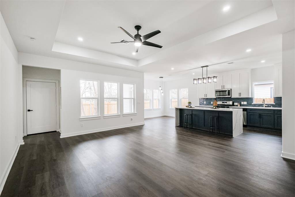 This modern kitchen features a spacious layout with a large marble island, elegant pendant lighting, and dark hardwood floors. It includes stainless steel appliances, white upper cabinets, and navy lower cabinets, complemented by a stylish dark blue backsplash. Large windows provide ample natural light.