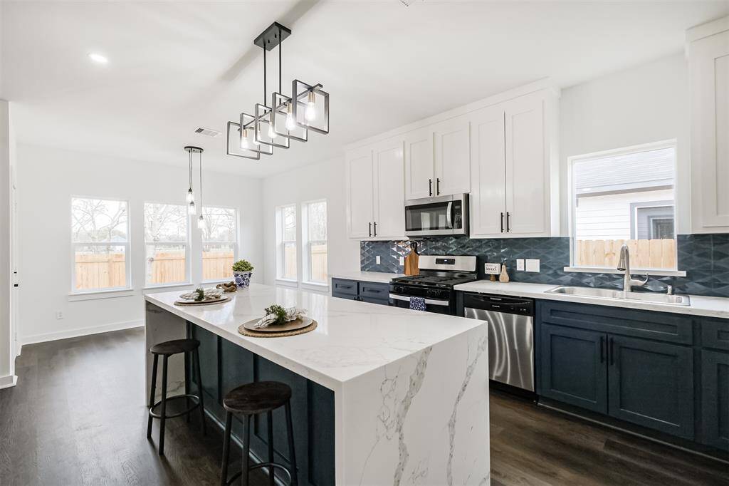 This photo showcases a modern open-concept kitchen and living area. It features sleek, dark wood flooring, a large island with marble countertops, navy blue lower cabinetry, and white upper cabinets. The kitchen is accented with a stylish backsplash and pendant lighting, creating a bright and inviting space.