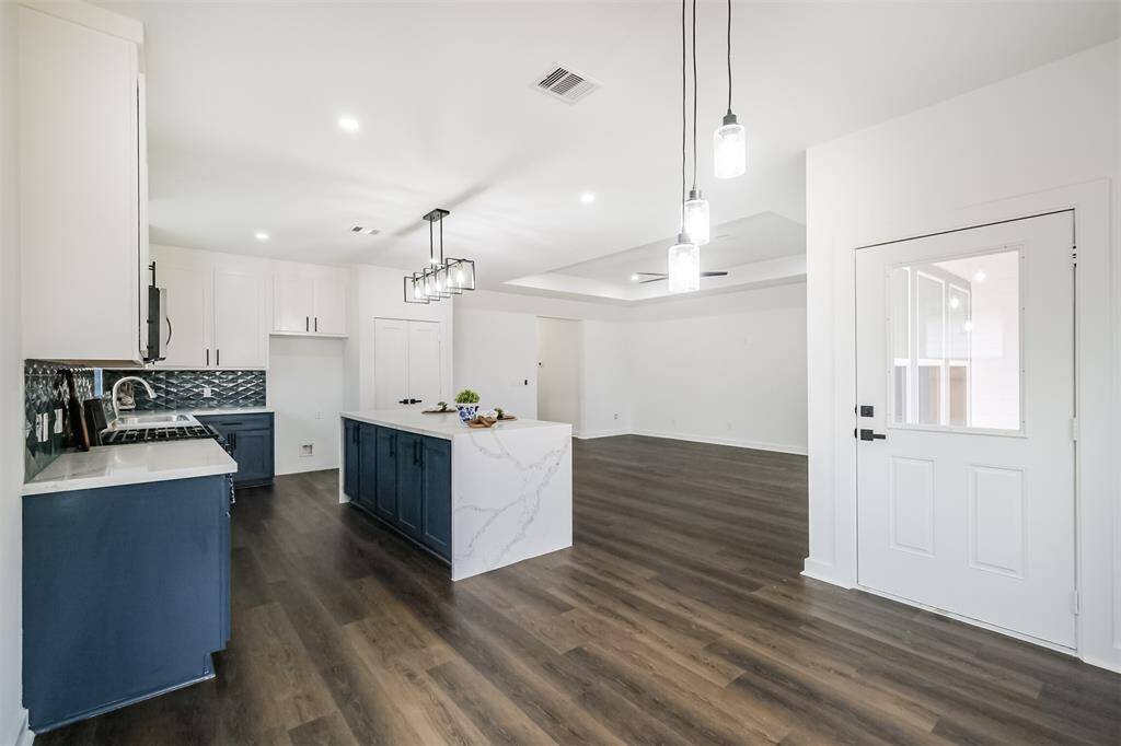 Bright, open-concept kitchen and living area with large windows, modern light fixtures, and dark wood flooring. Features a spacious island with a marble countertop and dark blue cabinetry.