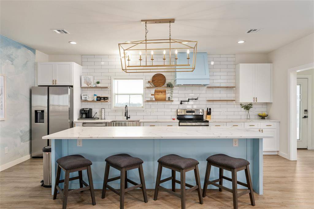 The large kitchen is open to the living room and features kitchen island accented in blue with gold stunning gold chandelier, stainless steel appliances, quartz countertops, tile running to the ceiling, open shelving, and a pot filler over the stove.