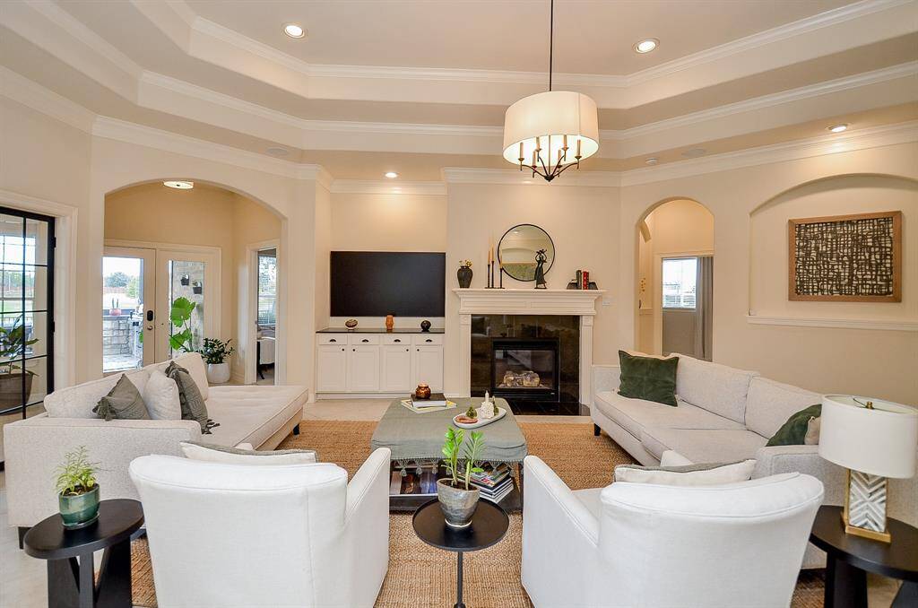A spacious, elegant living room featuring neutral colors, a cozy fireplace, and built-in cabinetry. The room is well-lit with a modern chandelier and recessed lighting. Large windows and an exit to the patio create a bright, inviting atmosphere.