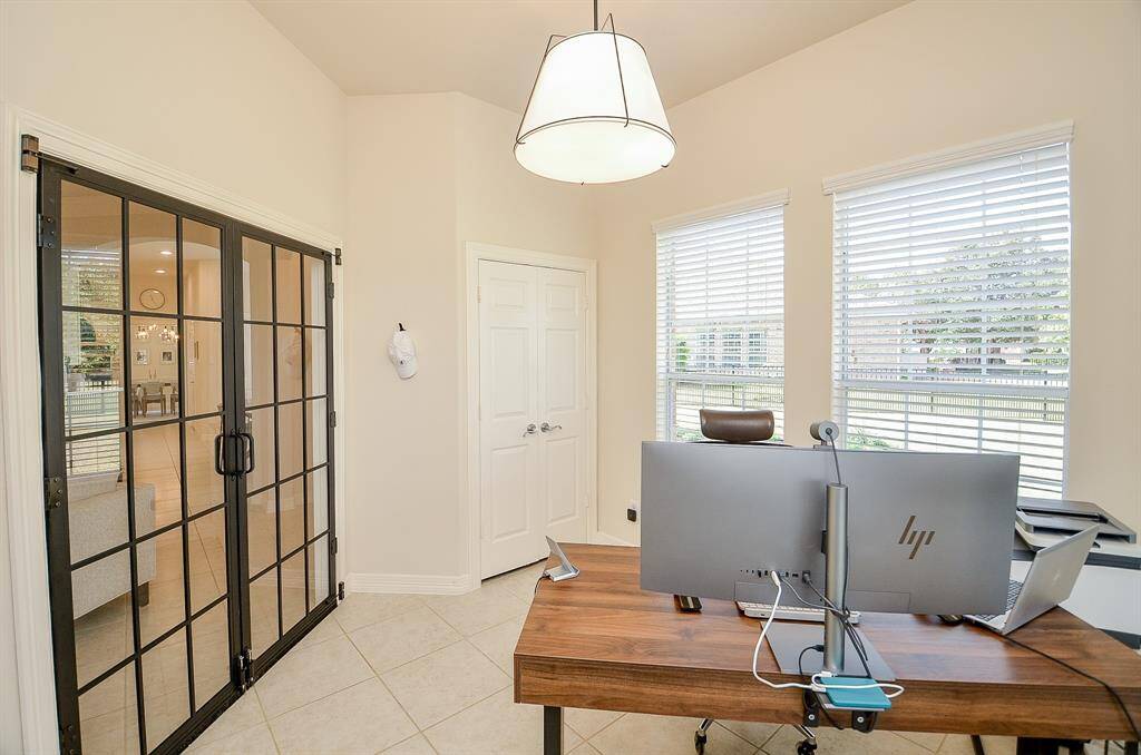 This bright home office features large windows with blinds, allowing natural light to flood the space. It includes modern glass double doors leading to a main living area, a sleek wooden desk, and contemporary lighting.