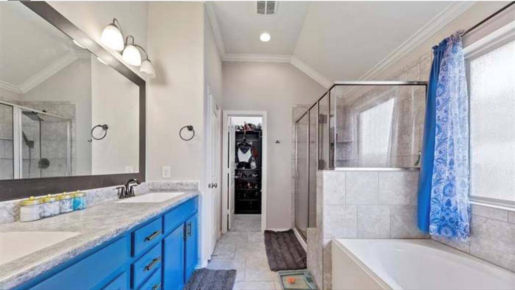 This bathroom features a double vanity with a striking blue cabinet, a large mirror, and overhead lighting. It includes a spacious tiled shower with glass doors and a separate bathtub. A window with blue patterned curtains allows natural light to fill the room, creating a bright and inviting space.