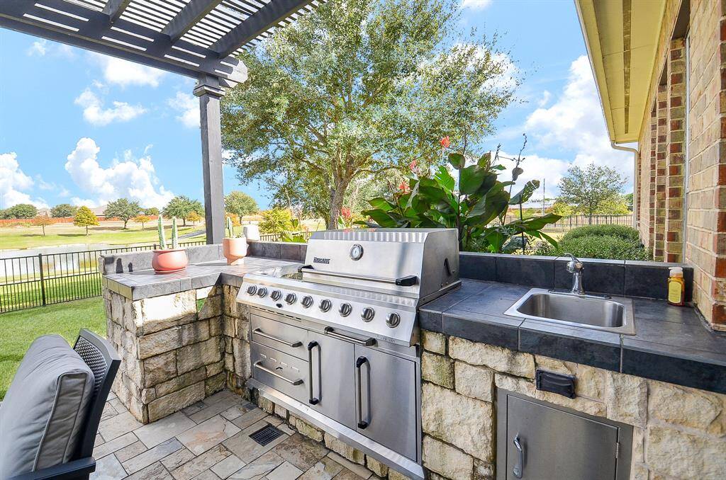 This inviting outdoor kitchen with a stainless steel grill, built-in stone countertops, and a sink. The space is covered by a pergola and overlooks a beautifully landscaped yard with a scenic view, perfect for entertaining.