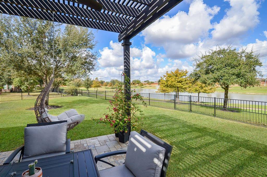 This photo showcases a serene backyard with a spacious patio, featuring comfortable seating under a pergola. The area overlooks a well-maintained lawn and a scenic pond, framed by mature trees and a fenced boundary, offering a peaceful and picturesque outdoor living space.
