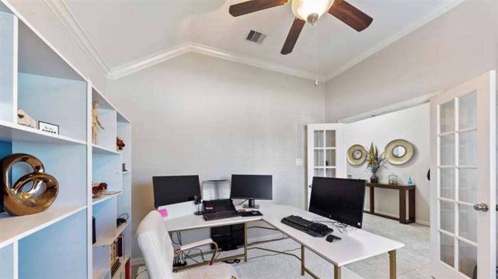 This photo showcases a bright home office with built-in shelving. The room has neutral walls, a ceiling fan, and French doors leading to a hallway.