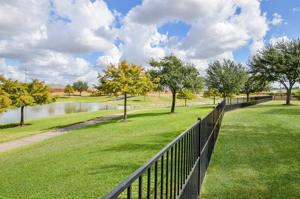 A serene, fenced backyard with lush green lawns and a paved walkway alongside a pond, offering scenic views and a peaceful atmosphere.