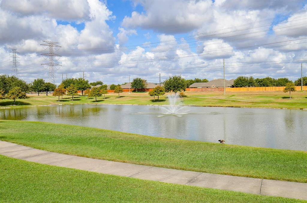 This photo showcases a serene residential area with a pond and fountain, surrounded by lush green lawns and trees. A sidewalk runs alongside the water, offering a peaceful setting perfect for leisurely walks.