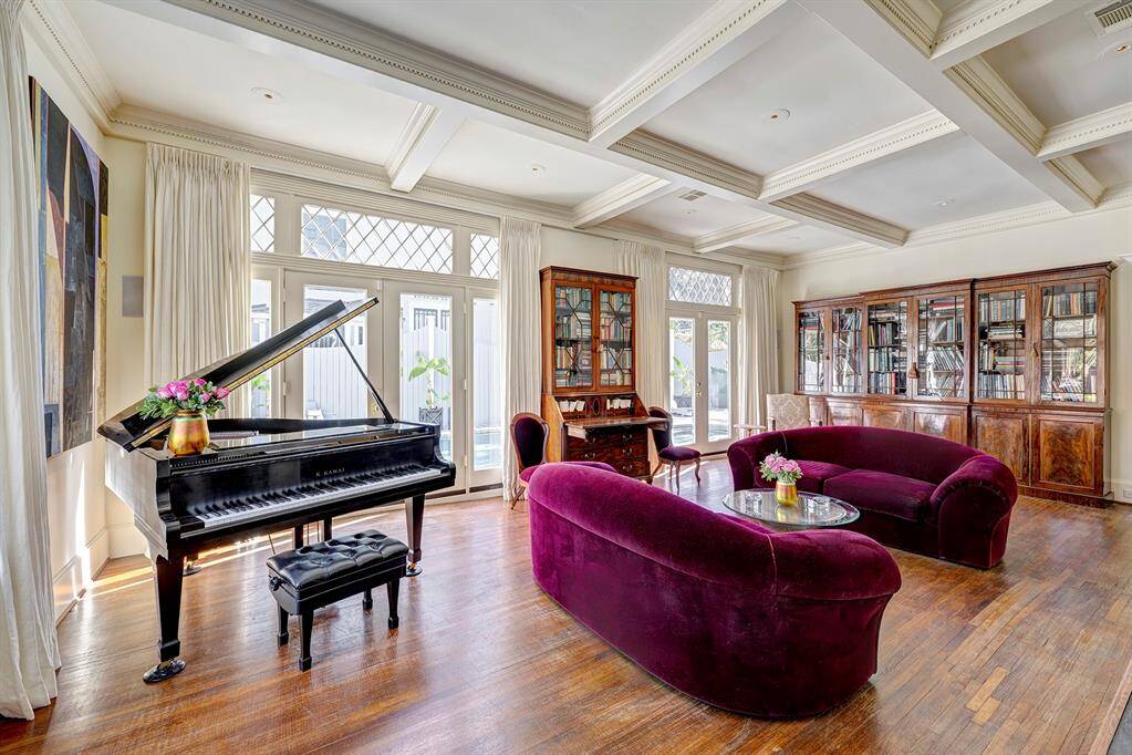 Stunning living room with a coffered ceiling and polished original wood floors that exude elegance. French doors open to a beautifully landscaped backyard with a sparkling swimming pool, seamlessly blending indoor and outdoor spaces. The soft sunlight enhances the natural tones, highlighting every intricate detail of the coffered ceiling and creating a warm, inviting, and sophisticated atmosphere.