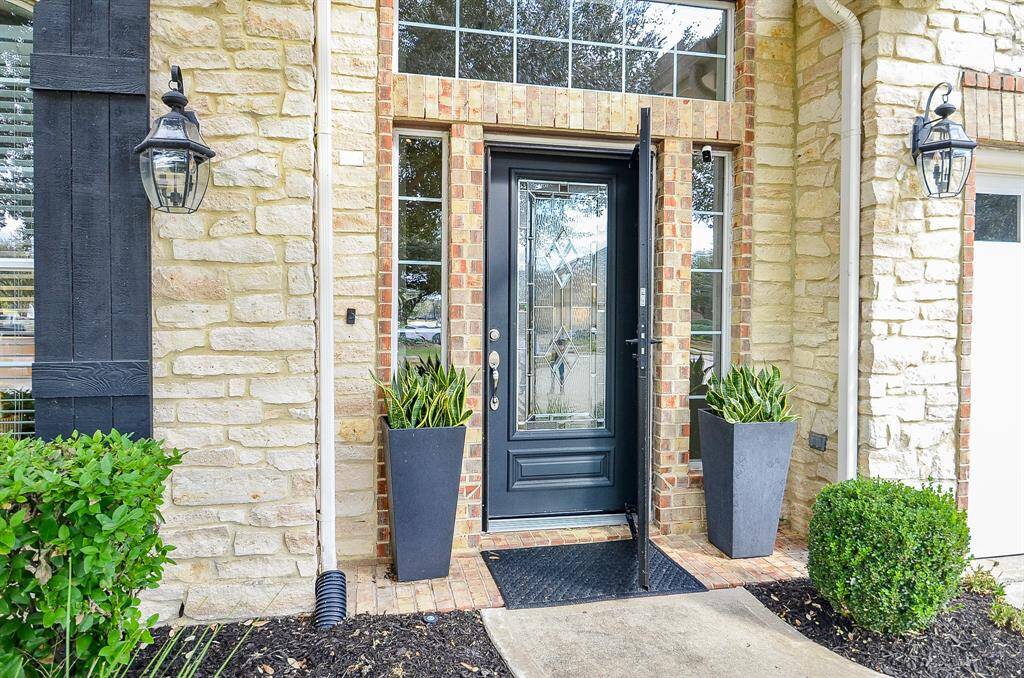 This photo showcases an elegant front entrance with a black door featuring decorative glass panels, framed by stone and brickwork. Flanked by modern planters and lantern-style lights, it offers a welcoming and stylish curb appeal.