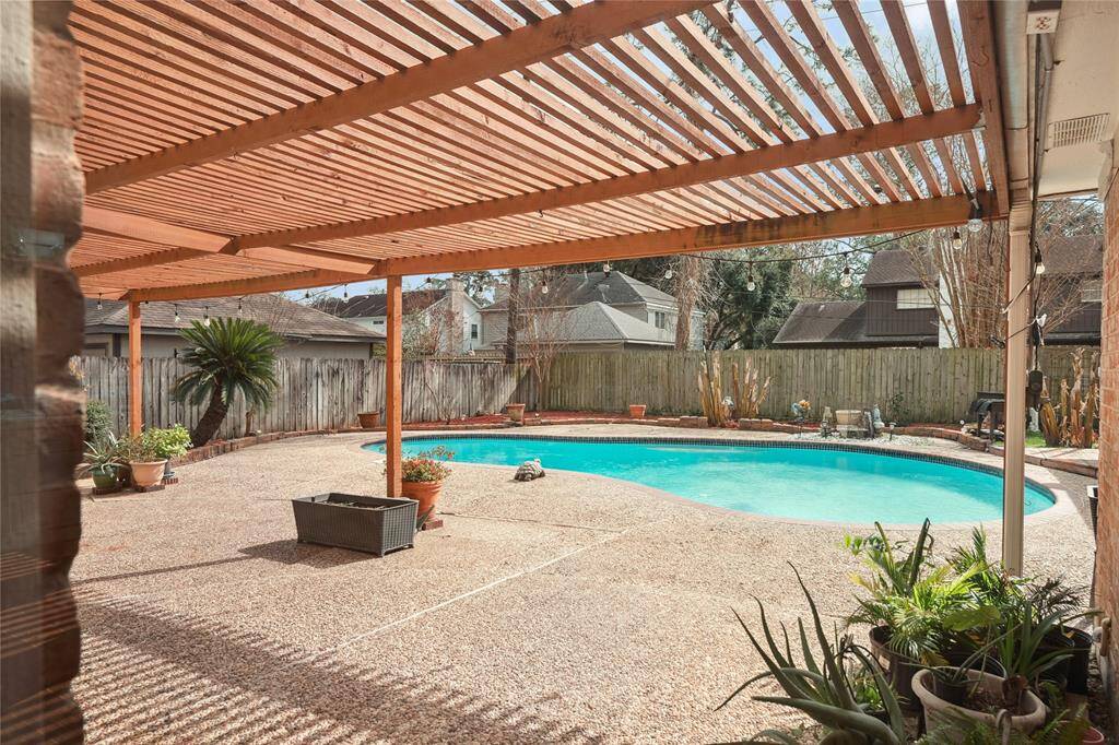 This backyard features a spacious pool with a surrounding patio area, partially covered by a wooden pergola.
