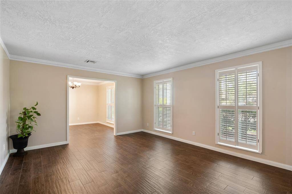 This spacious, light-filled living room features rich wood-look tile flooring, neutral walls, and large windows with plantation shutters. A charming dining area is visible through an open doorway.
