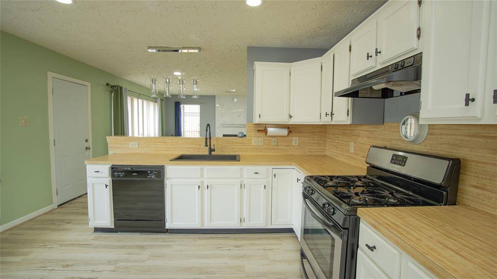 This kitchen features white cabinetry, a black stove and dishwasher, laminate-wood countertops, and a light green accent wall. It opens into a spacious living room with plenty of natural light.