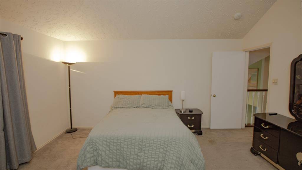 This secondary bedroom features a cozy layout with a bed positioned against a light painted wall. It includes bedside tables and a standing lamp for lighting. The room has carpeted flooring, neutral decor, and an adjoining doorway.