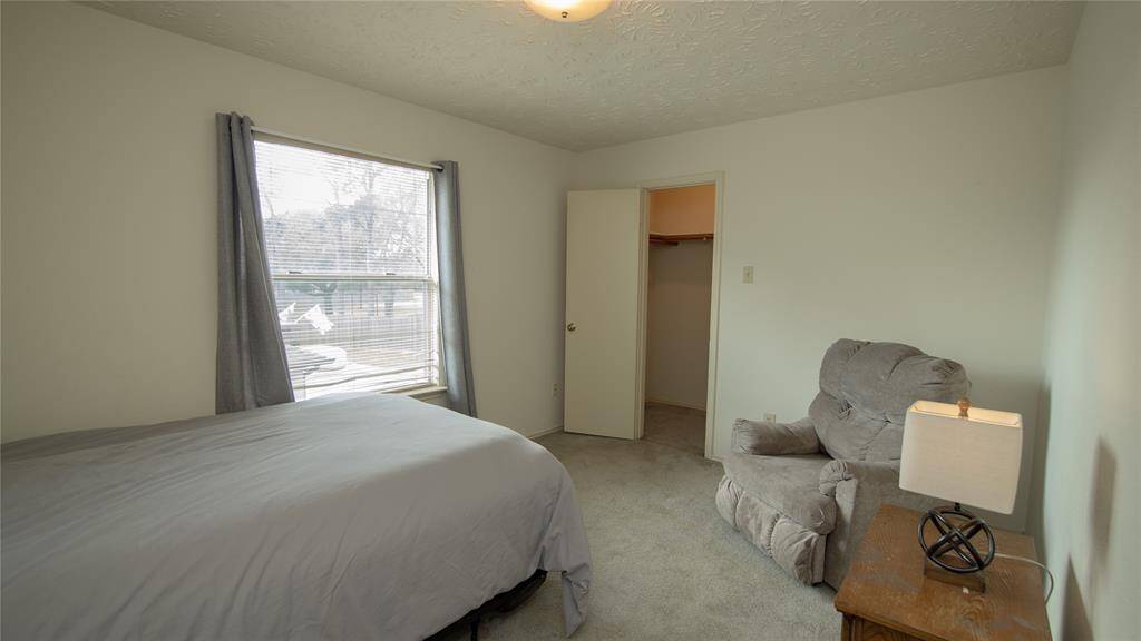 The secondary bedroom features a large window with natural light, a comfortable bed, a plush armchair, and a small wooden side table with a lamp. A closet is accessible through an open door, adding practical storage space.