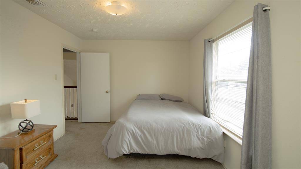The secondary bedroom features a large window with natural light, a comfortable bed, a plush armchair, and a small wooden side table with a lamp. A closet is accessible through an open door, adding practical storage space.