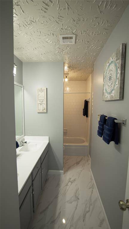 The secondary bathroom features a modern design with light gray walls and a textured ceiling. It has a white double vanity with ample counter space, a bathtub with a white tile surround, and elegant marble-like flooring.