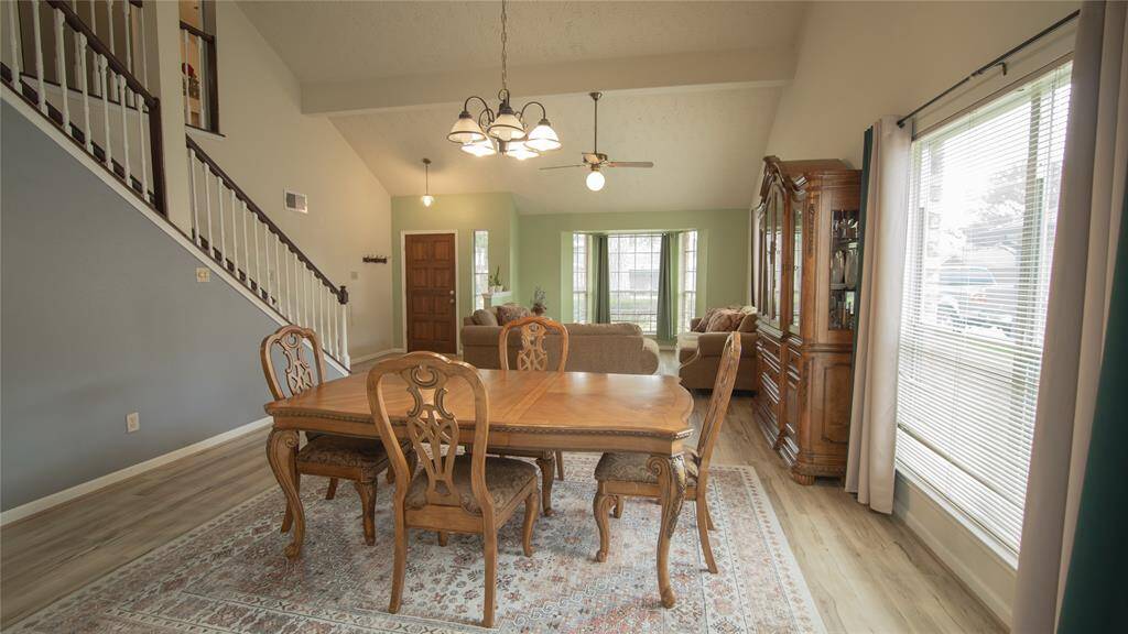 This inviting living space features an open floor plan with a dining area, high ceilings, and large windows for natural light. The room is accented by a wooden staircase and a classic chandelier, offering a warm and elegant atmosphere.