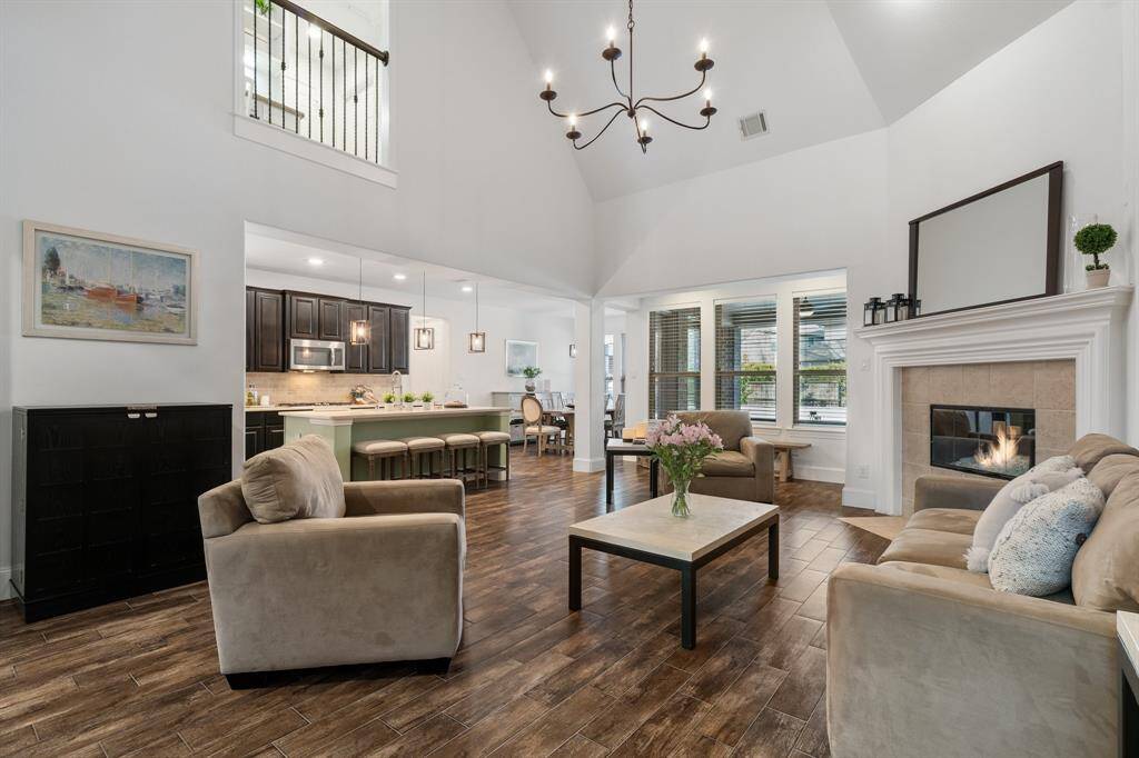 This inviting living area features high vaulted ceilings, a cozy fireplace, and elegant wood like tile flooring. It seamlessly connects to an open kitchen with dark cabinetry and a spacious island with upgraded travertine counters providing a perfect space for entertaining. Large windows across the back offer plenty of natural light.
