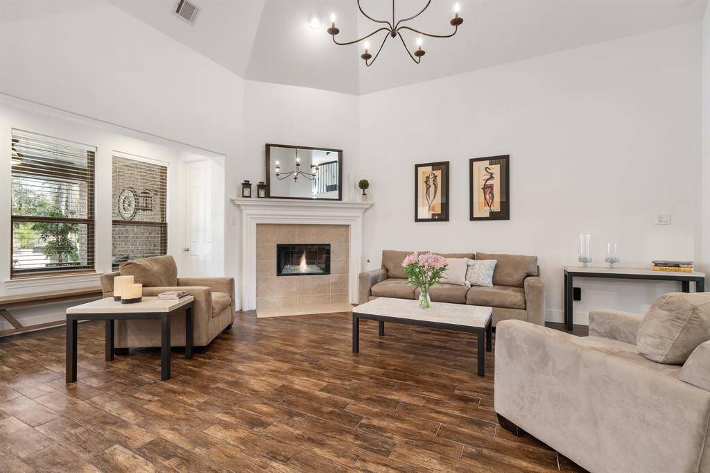 This inviting living room features a cozy fireplace, elegant chandelier, and warm wood like tile looring. Neutral-toned furniture and tasteful wall art create a welcoming ambiance. Large windows provide ample natural light, enhancing the room's spacious feel.