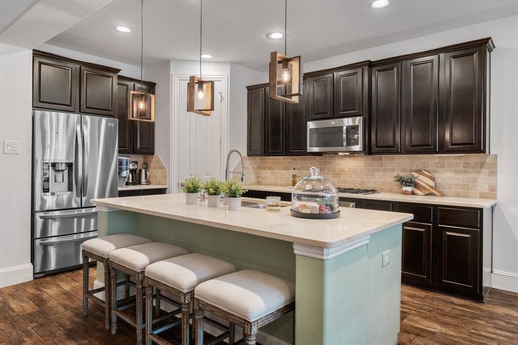 This kitchen features dark wood cabinets, stainless steel appliances, and a large island with ample room for seating. The space is well-lit with pendant lights and has a warm, inviting atmosphere with a stylish backsplash and wood looking tile floors.