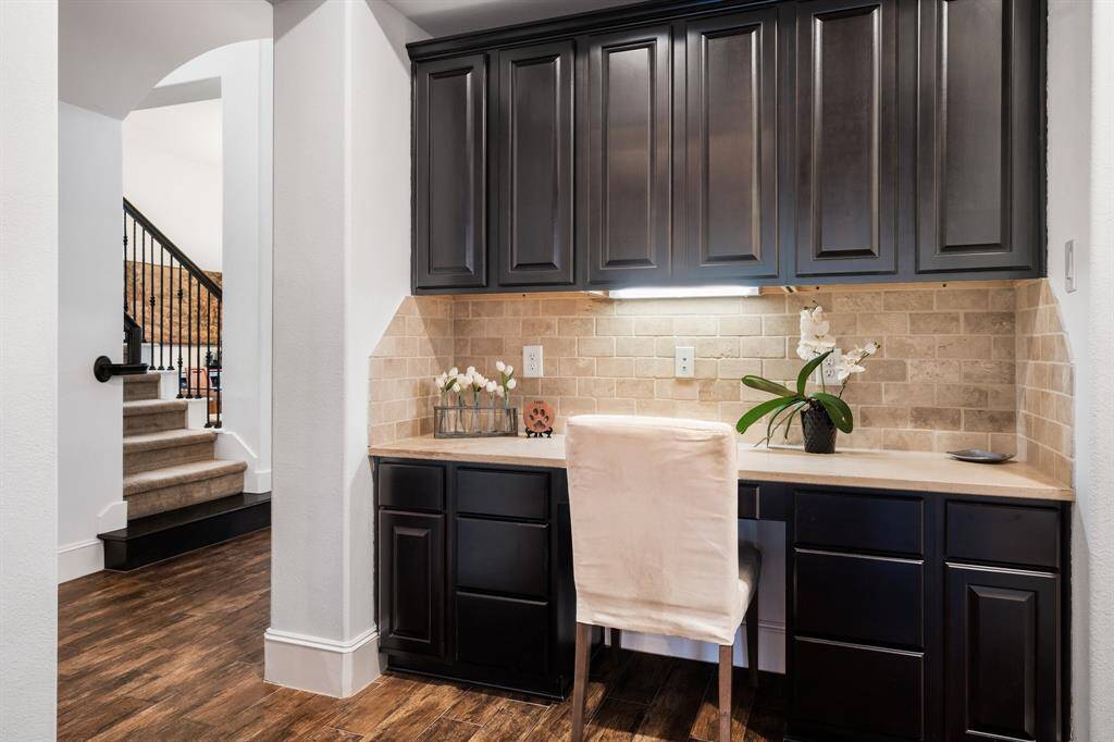 This photo shows a built-in desk area with elegant dark cabinetry and a beige stone backsplash. The space is well-lit and adjacent to a stairway with a wooden and carpet finish, providing a cozy and functional workspace within the home.
