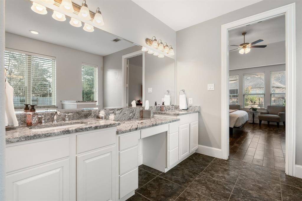This photo showcases a modern bathroom with a dual-sink vanity featuring granite countertops and ample storage. It opens into a spacious bedroom with large windows, allowing for plenty of natural light. The space is well-lit and has a clean, contemporary design.