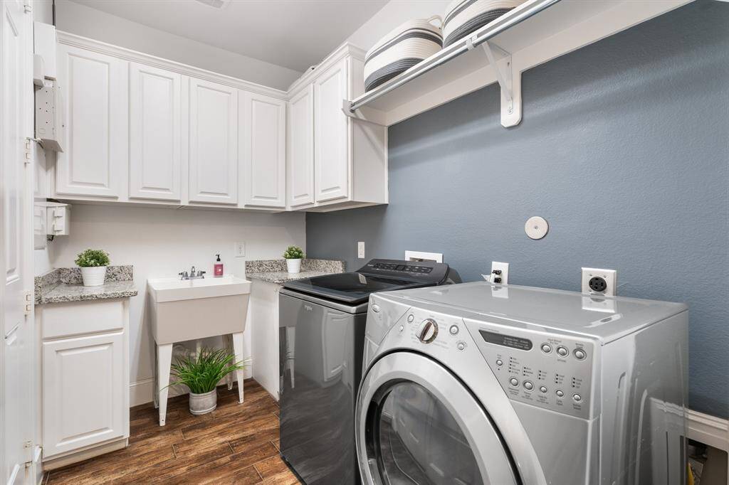 This photo shows a modern laundry room featuring white cabinetry, a utility sink, and a washer and dryer set. The room has wood-style tile flooring and a calming blue-gray wall, providing a clean and functional space for laundry tasks.