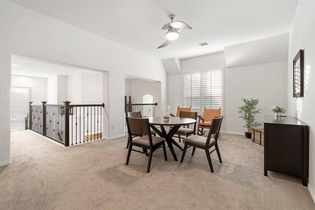 The upstairs game room is a bright, open space with neutral carpet and walls, featuring a spacious area for all your games with a modern ceiling fan and plantation wooden window shutters. It's open loft is surrounded with black iron railing overlooking the floor below, adding a sense of openness and connection between levels.