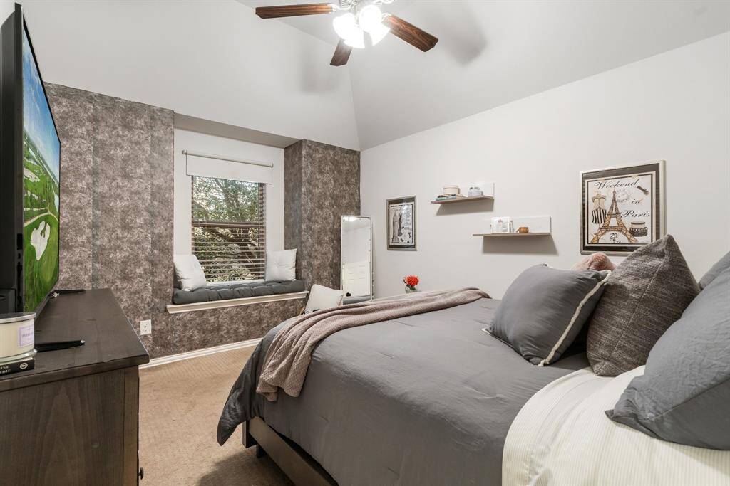 This cozy bedroom features a vaulted ceiling with a fan, and a window nook with seating. The room is decorated with modern wallpaper offering a comfortable and stylish retreat.