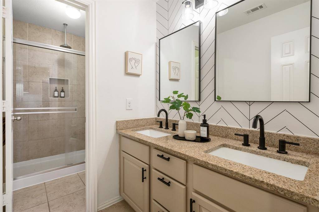 This updated bathroom features a modern design with a double vanity, granite countertops, and black fixtures. There's a large glass-enclosed shower with beige tile, and the walls have a stylish chevron pattern.