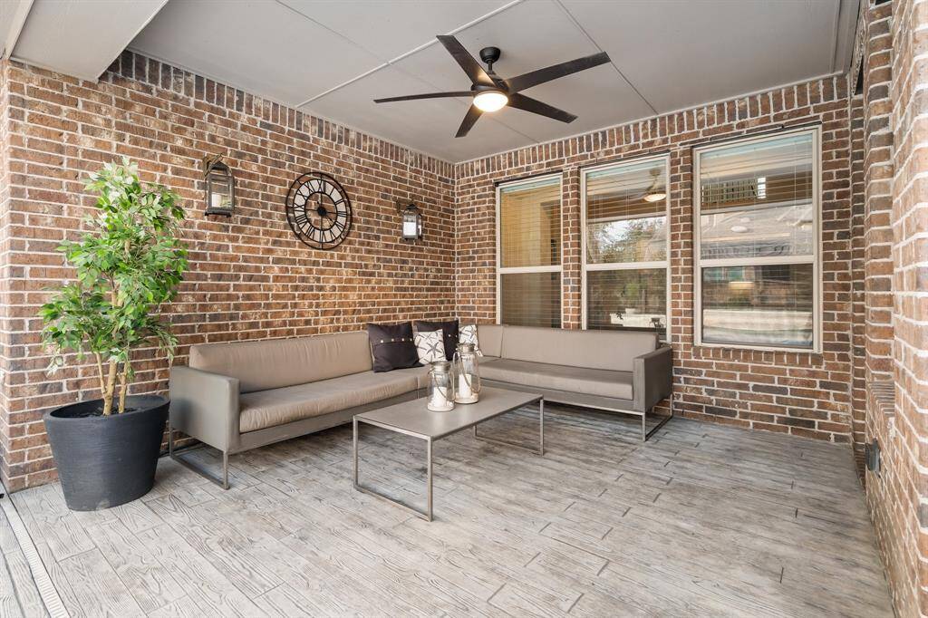 This inviting outdoor patio features a cozy seating area with modern furniture against a stylish brick wall. It includes a ceiling fan, ambient lighting, and decorative elements, perfect for relaxation or entertaining.