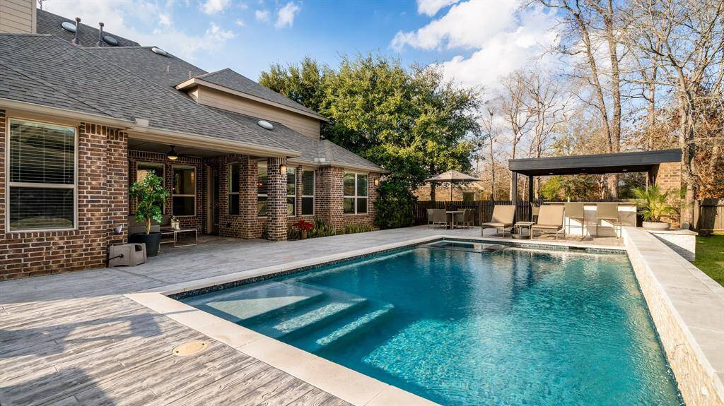 This photo showcases a backyard with a spacious in-ground modern pool with serene waterfall flowing from the retaining wall and spa surrounded by a covered patio. The brick home features large windows and a covered patio area. There's a pergola with seating and lush greenery, providing a serene outdoor living space.